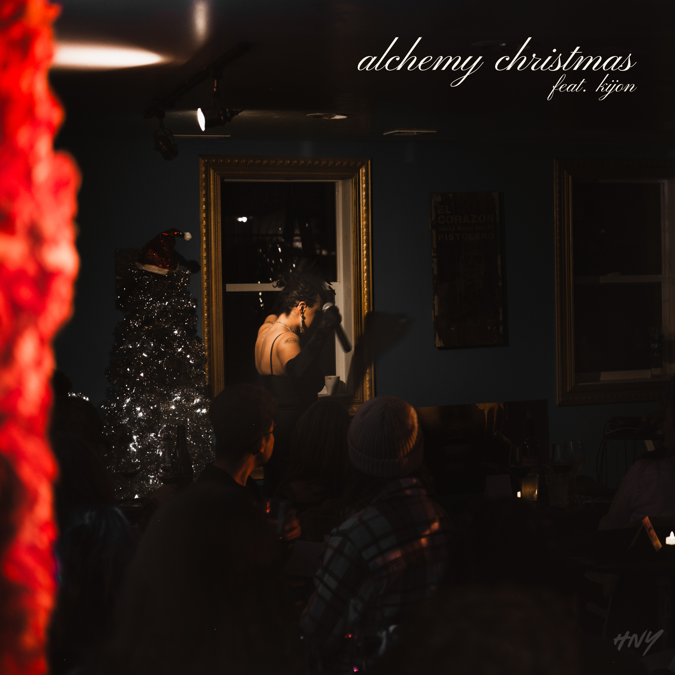HNY performs on stage with a microphone in a dark room decorated for Christmas, with a Christmas tree and a gold-framed mirror in the background, and a small audience watching.