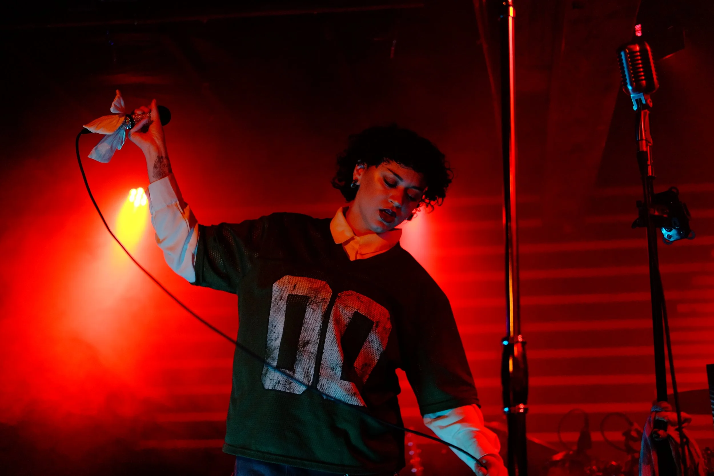 HNY with curly hair singing into a microphone on stage at Sunset Tavern with red and yellow lighting.