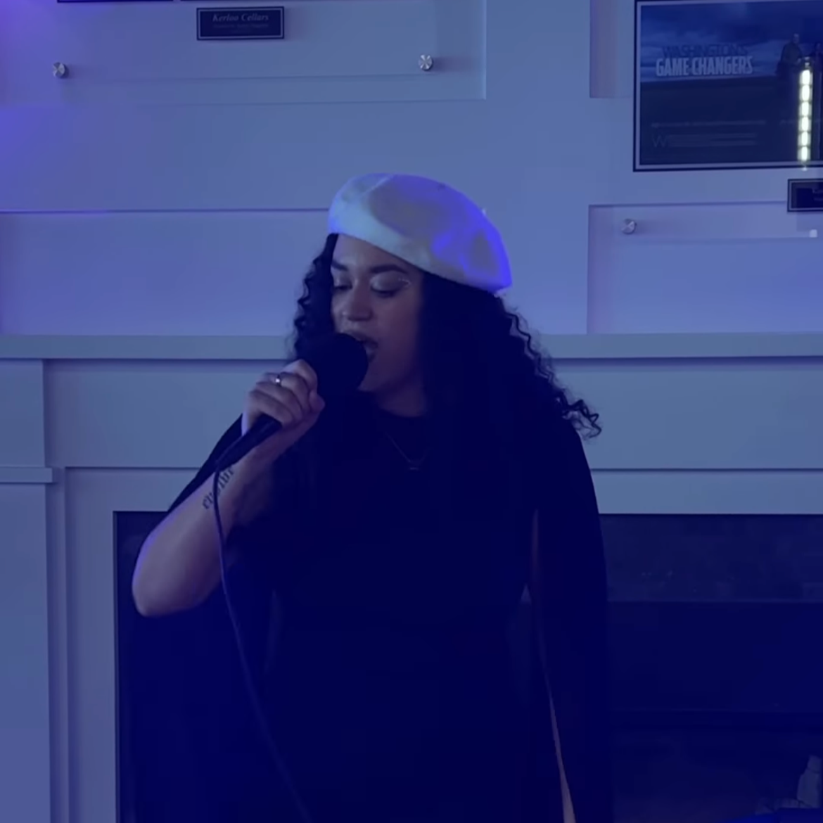 HNY with curly black hair wearing a white beret sings into a microphone in a dimly lit room.