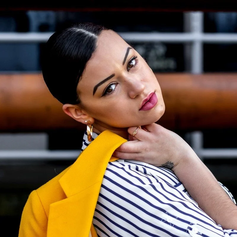 HNY with dark hair, bold eyebrows, and pink lips wearing a striped shirt and a yellow blazer, resting her chin on her hand, looking at the camera.
