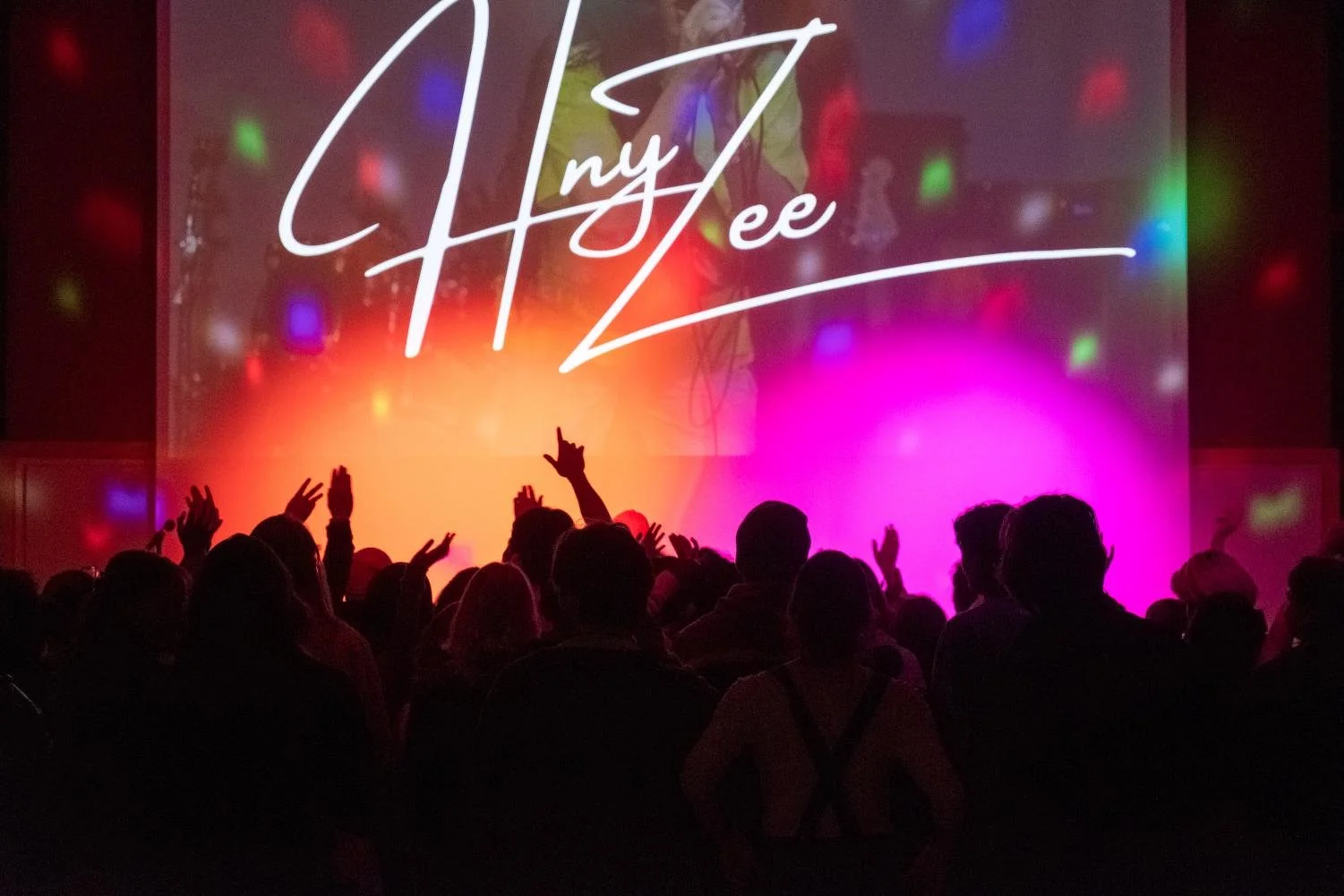 A crowd of people at Whitman College with colorful lights and a large screen display that reads 'HNYZEE' in stylized font.