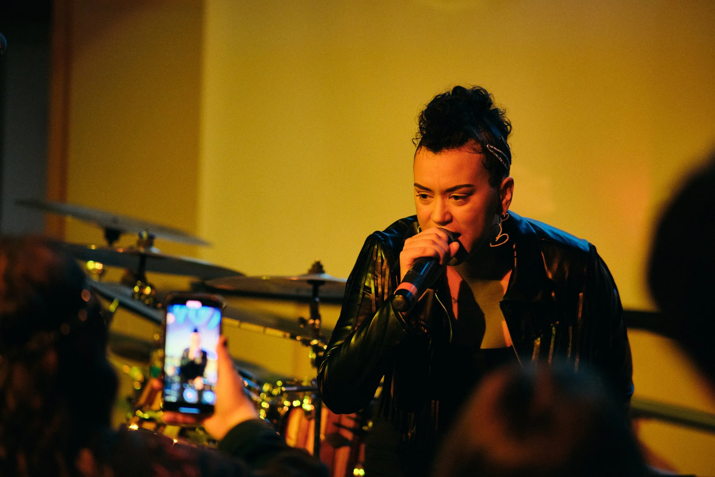 HNY performing on stage at Whitman College with a microphone, wearing a black leather jacket and heart-shaped earrings, with a yellow background and drums in the background.