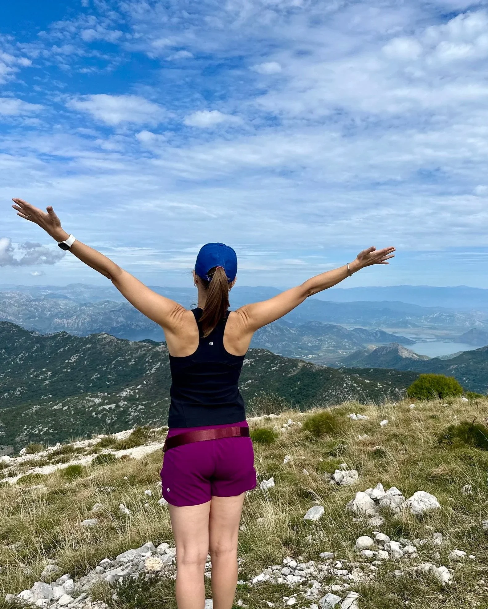 A woman in athletic clothing with arms raised standing on a mountain overlooking a landscape of mountains and a lake.