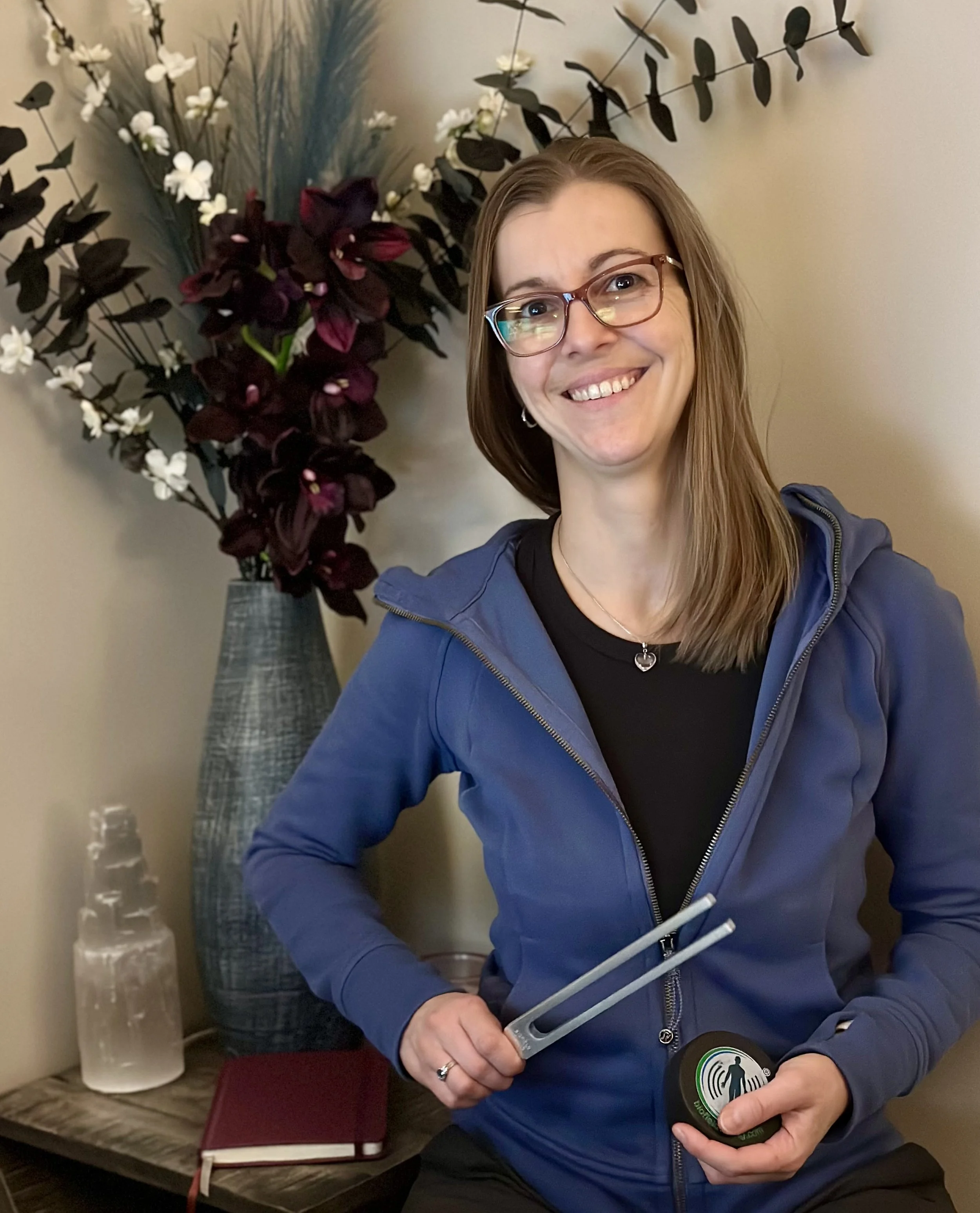 A smiling woman with glasses and long brown hair in a blue jacket holding a silver tool and a round black device with a green speaker icon. Behind her, there is a large flower arrangement with dark purple and white flowers in a tall vase and a small clear glass container on a wooden table.