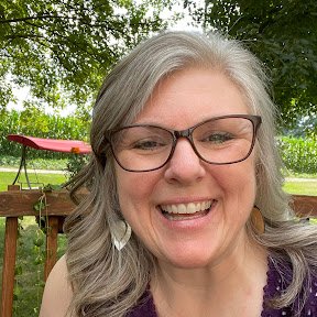 Close-up of a smiling woman with gray hair and glasses, outdoors in a park with trees and a wooden bridge in the background.