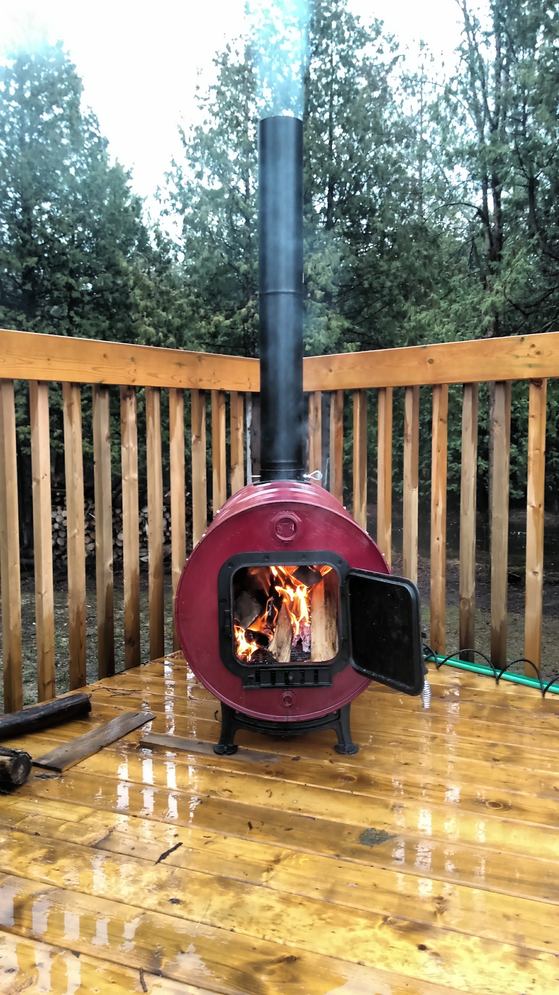A small red wood stove with an open door, burning logs inside, on a wet wooden patio with a natural wood railing, surrounded by trees.