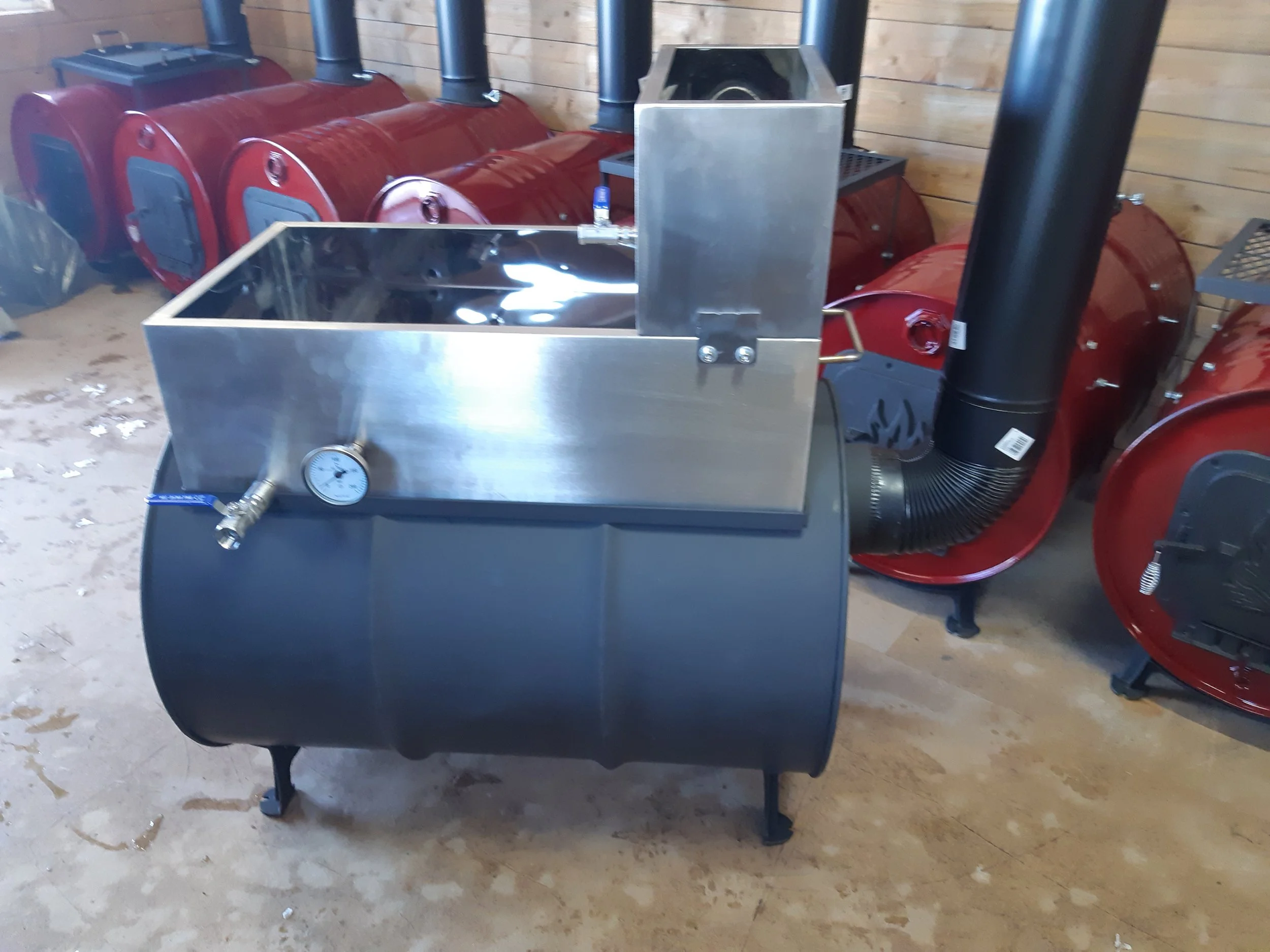 A large stainless steel hot liquor tank with a temperature gauge and tap, connected to a black industrial duct, in a brewery equipment storage area with red fermenters or brewing tanks in the background.