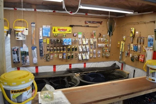A workshop with a pegboard wall displaying various tools and supplies, including paints, brushes, and hardware. There are yellow buckets on the counter and a workbench in the foreground.