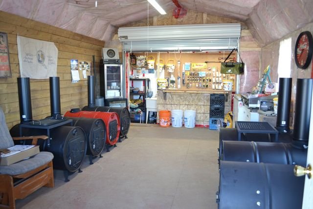 Workshop with various wood and metal tools, a workbench, and large black and red wood stoves, along with storage shelves and a clock on the wall.