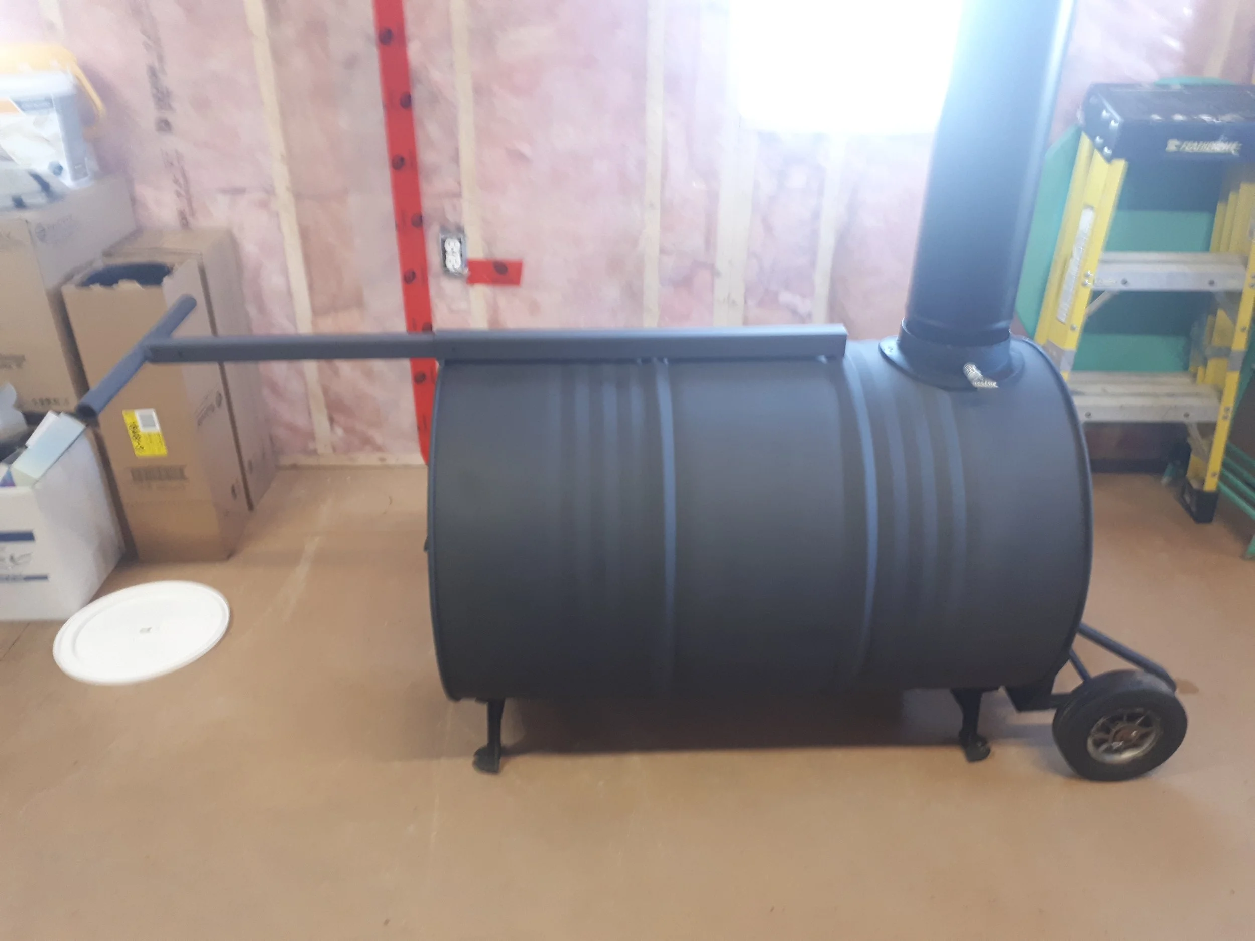 A black barrel smoker with a chimney pipe in a garage or storage room, with boxes and a ladder in the background.