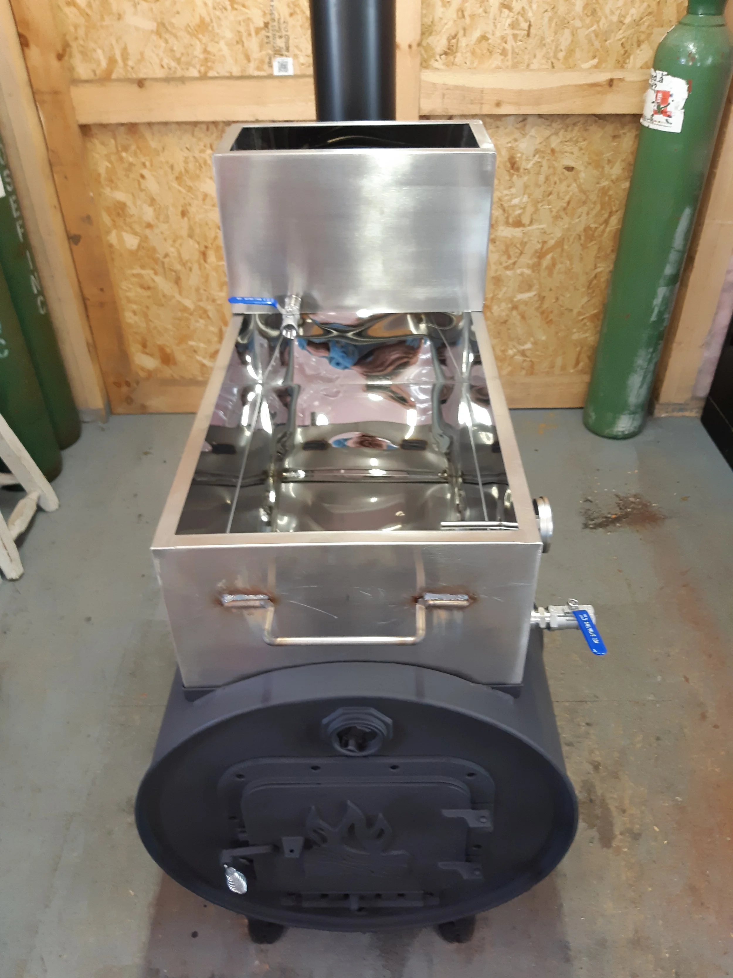 Stainless steel portable stove with a water tank and faucet, placed on a wheeled cart in a workshop. Green compressed gas cylinders are visible on the sides.