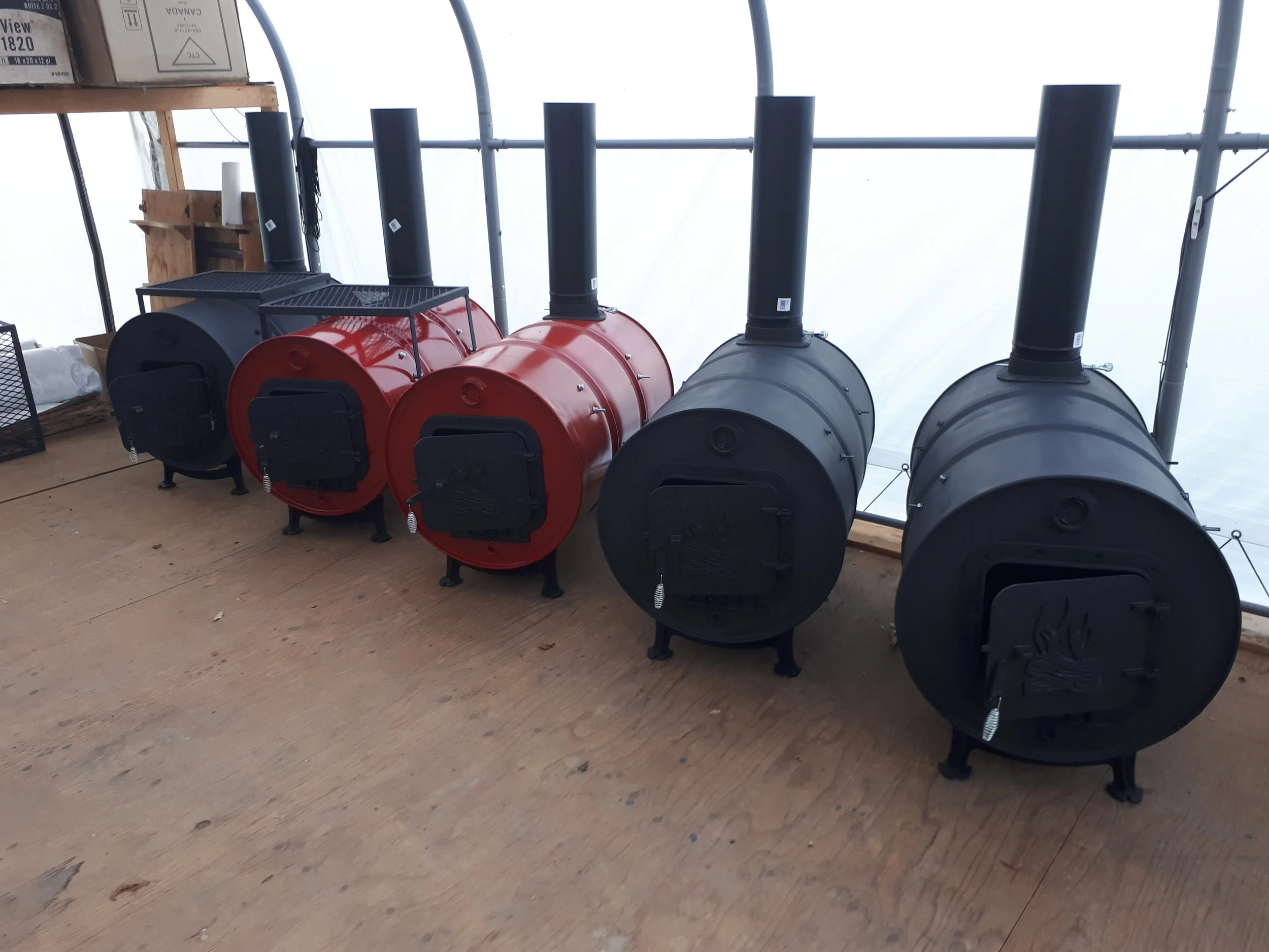 Four small wood-burning stoves, two red and two black, lined up on a wooden floor inside a greenhouse or shed.
