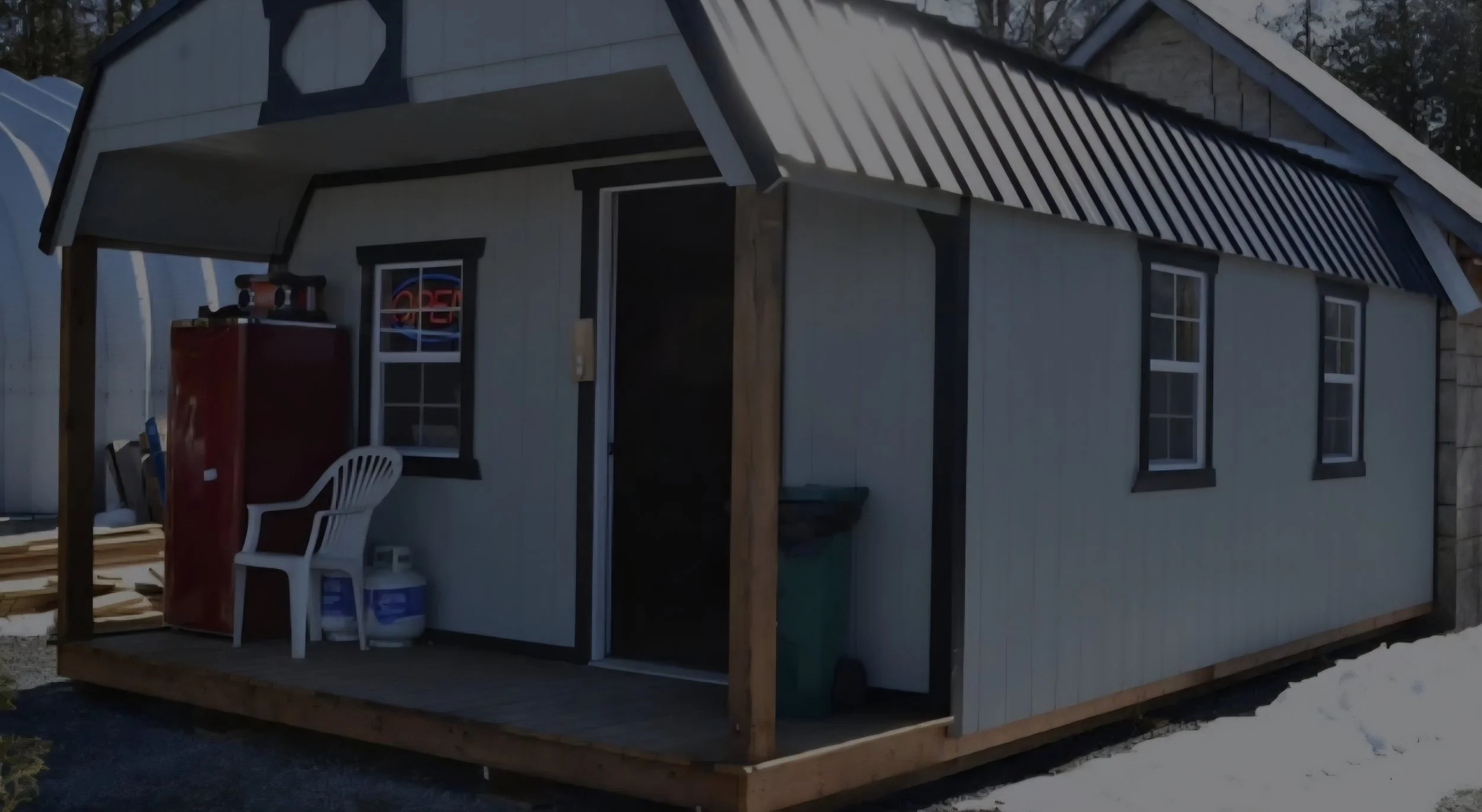 Small shed with a porch, two windows, and a door, situated on a snowy ground, with a red refrigerator, patio chair, and propane tanks on the porch.