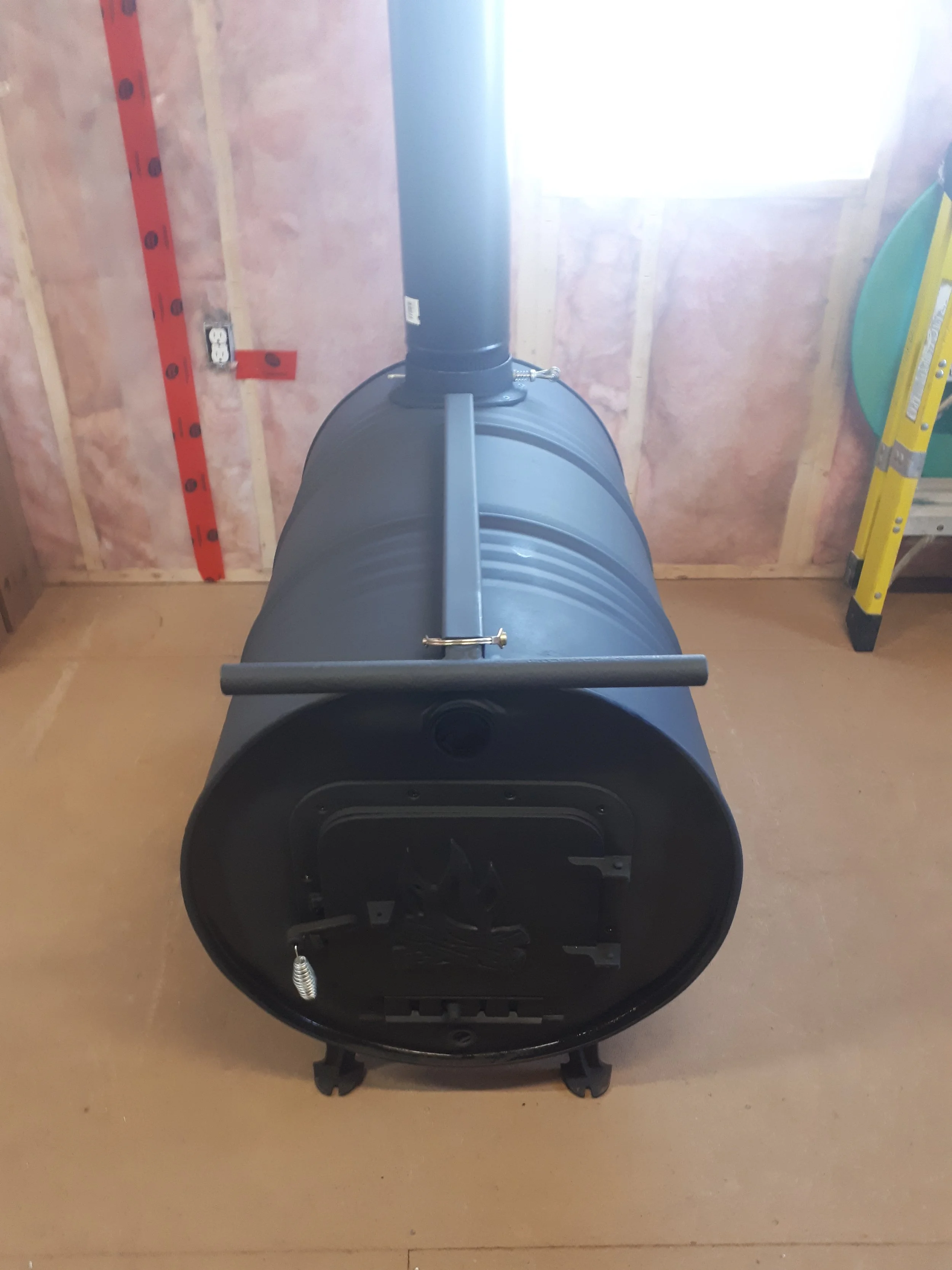 Black pellet stove inside a room with unfinished walls, a window, a yellow ladder, and a red measuring guide on the wall.