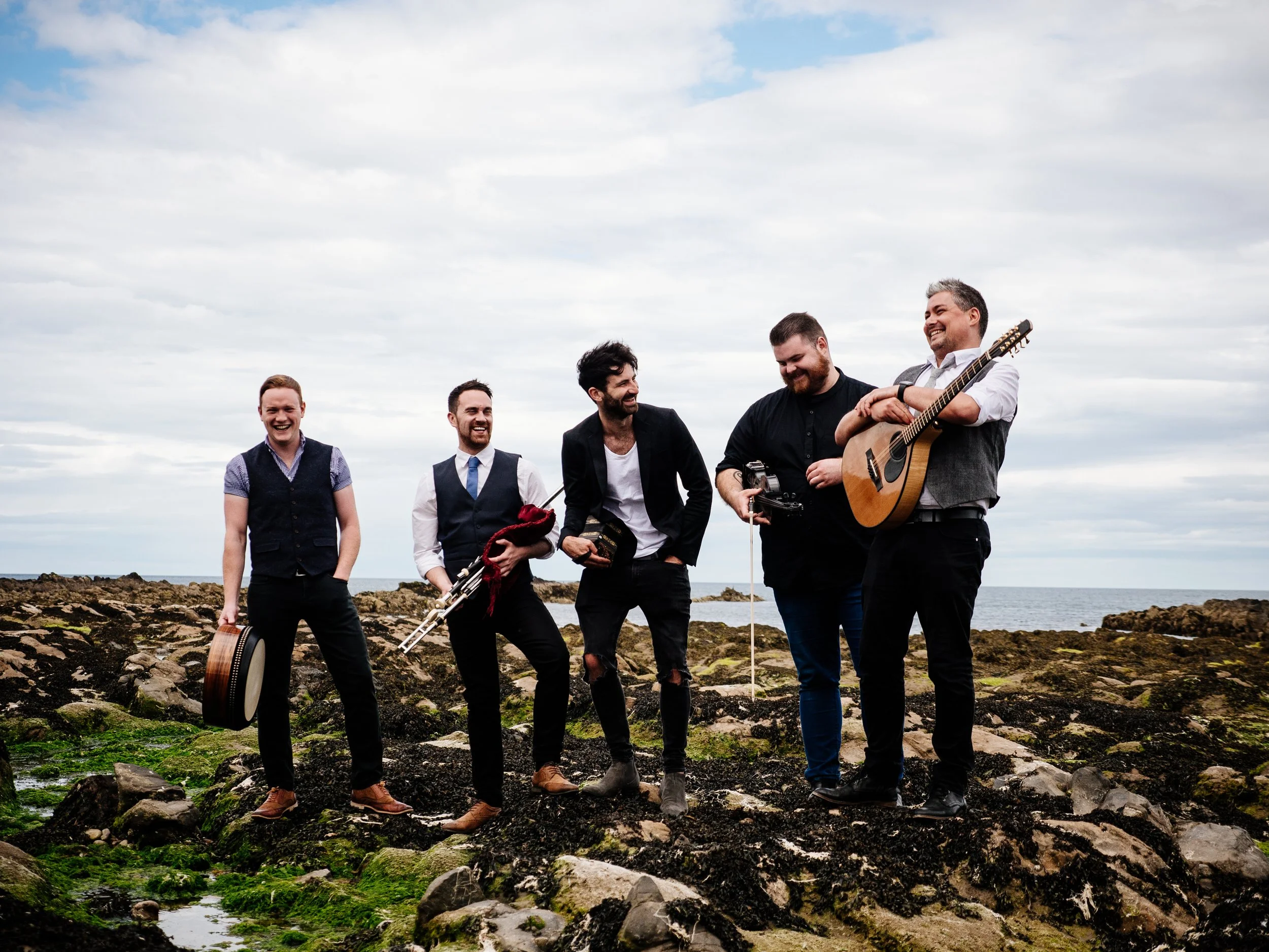 Five men standing on rocky coast with ocean in background, holding musical instruments including a drum, flute, and guitar, smiling and laughing.
