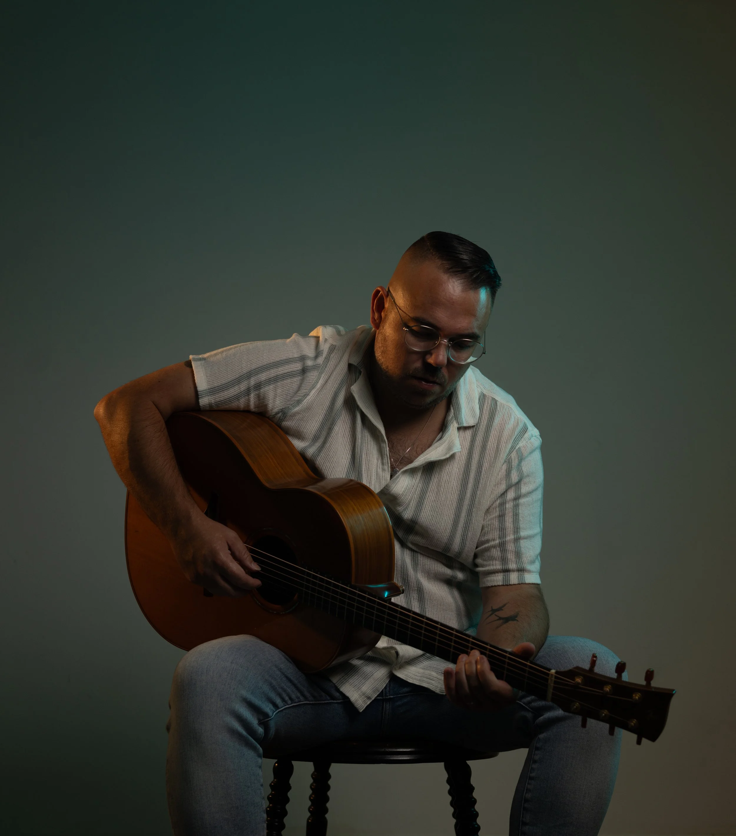 A man sitting on a chair playing an acoustic guitar in a dimly lit studio with a dark green background.