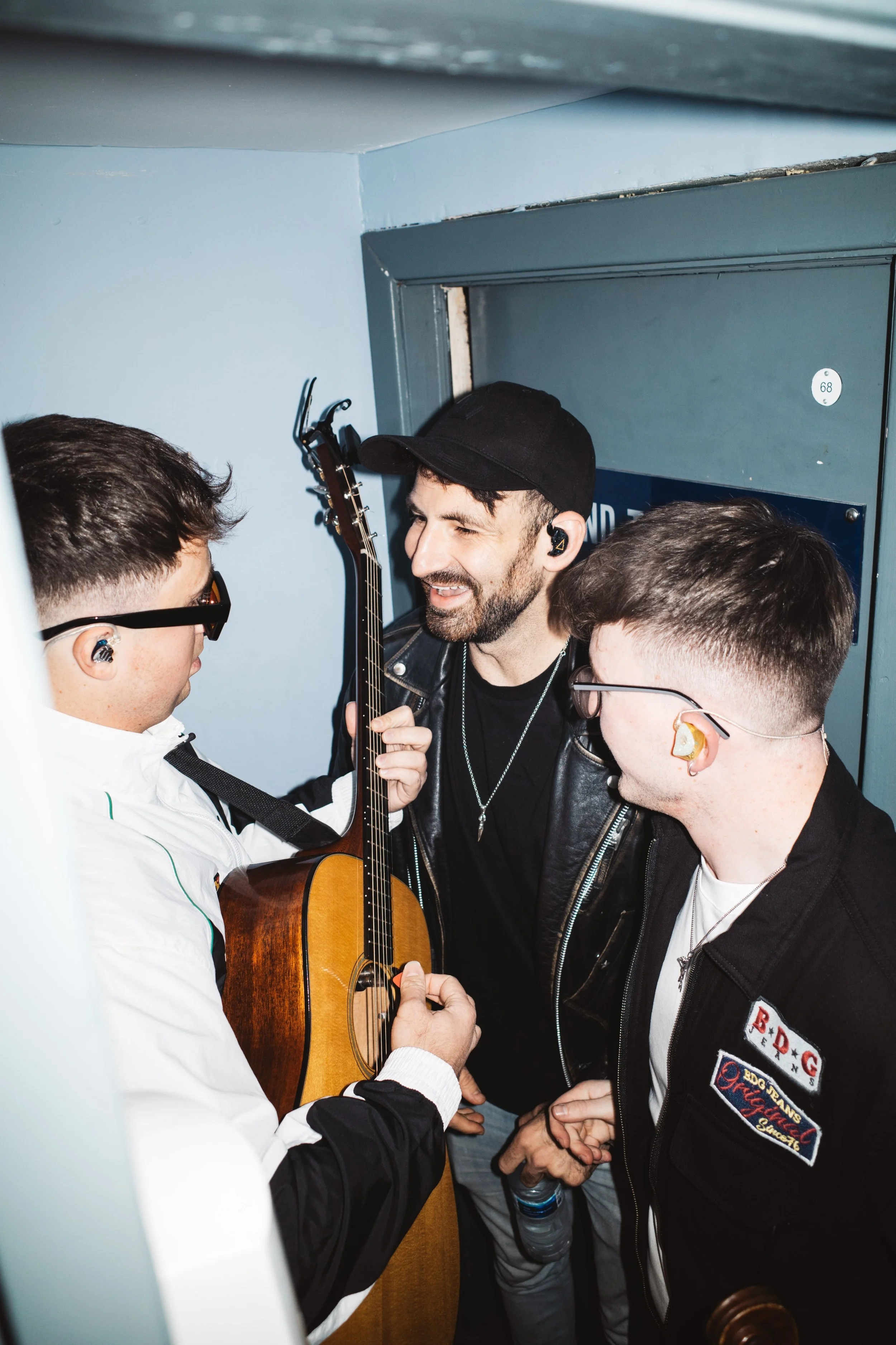 Three young men in a narrow, enclosed space, possibly behind the stage, are smiling and engaging with each other. One of them is holding an acoustic guitar, while two are wearing in-ear monitors and casual streetwear.