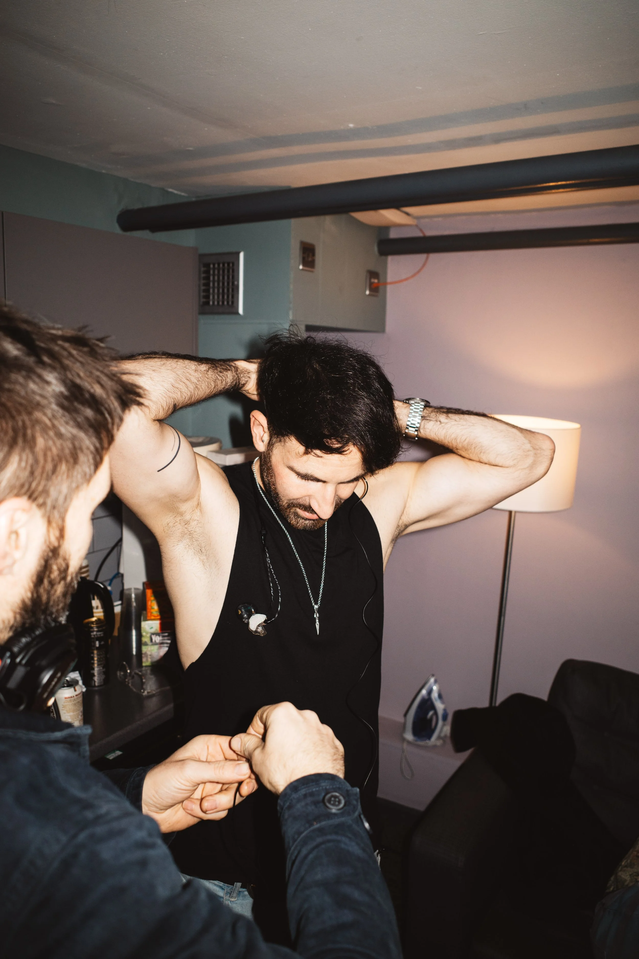 A man with dark hair and a black sleeveless shirt is adjusting his hair, while another man with a beard and a black jacket is holding his hands and assisting. The scene appears to be indoors, in a room with a lamp, an ironing board, and various personal items.