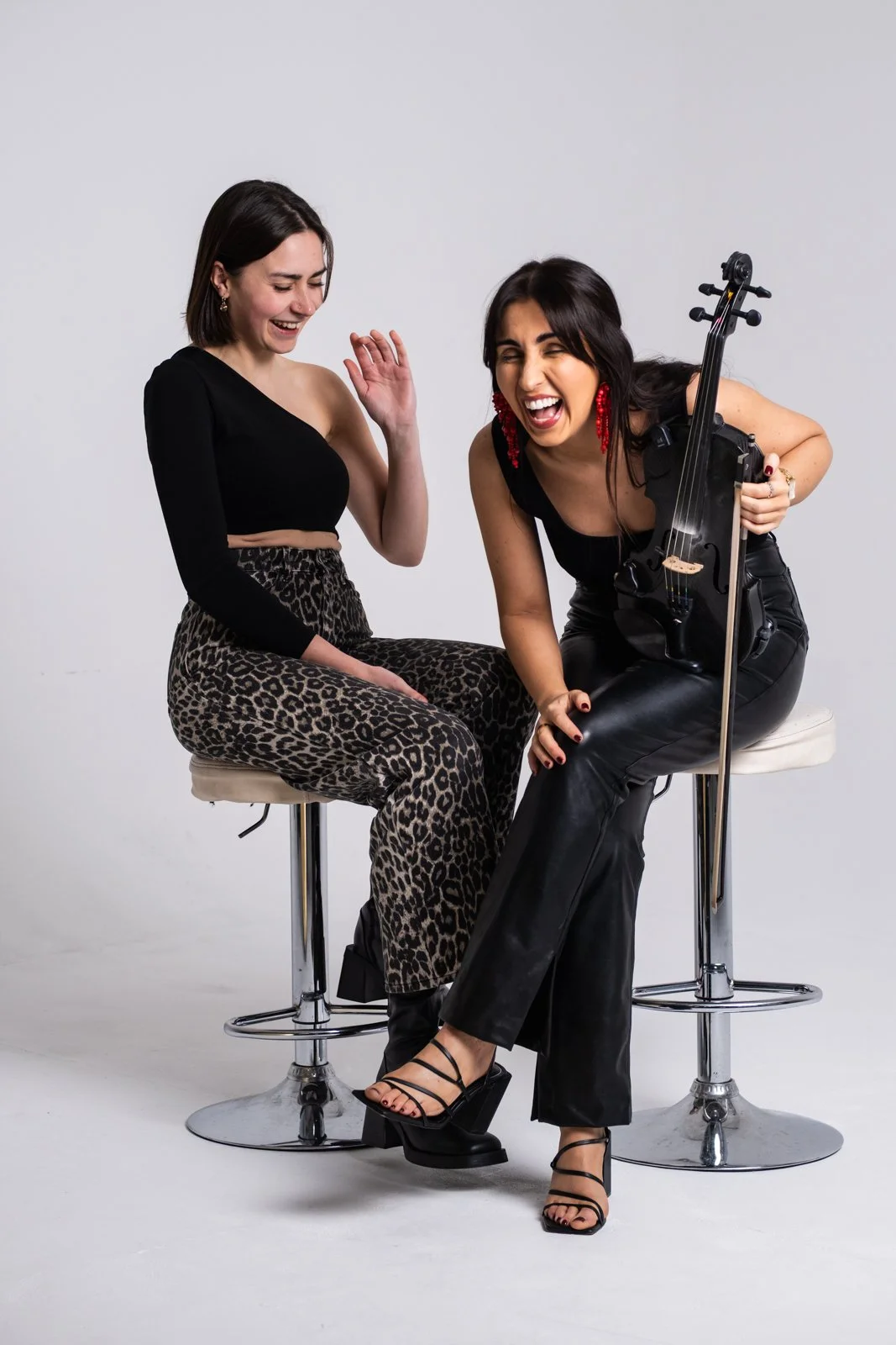 Two women sitting on high seats, laughing and enjoying themselves. One woman holds a violin, wearing black leather pants and black high heels, with red earrings. The other woman is wearing leopard-print pants and a black off-shoulder top. They are in a neutral studio setting.