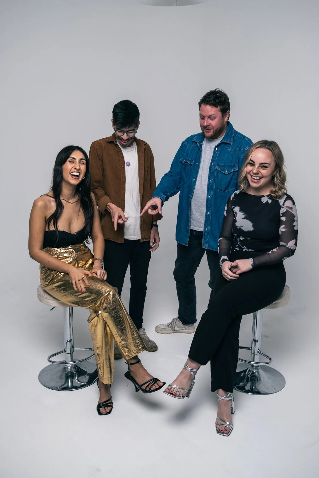 Four friends laughing and having fun in a photo studio with a white background, two women sitting on stools and two men standing next to them.