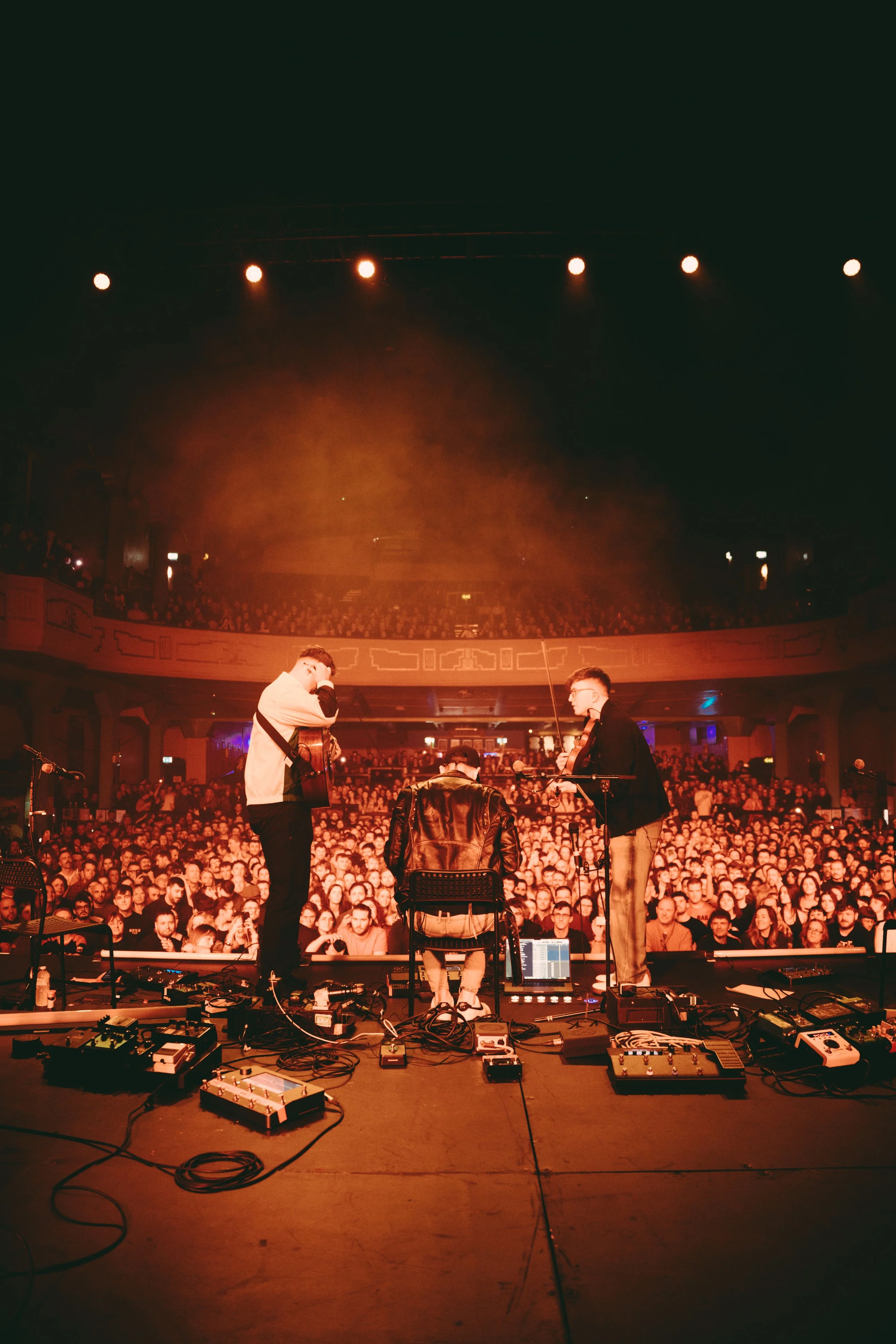 Band performing on stage with musical equipment in front of a large audience in a concert hall.