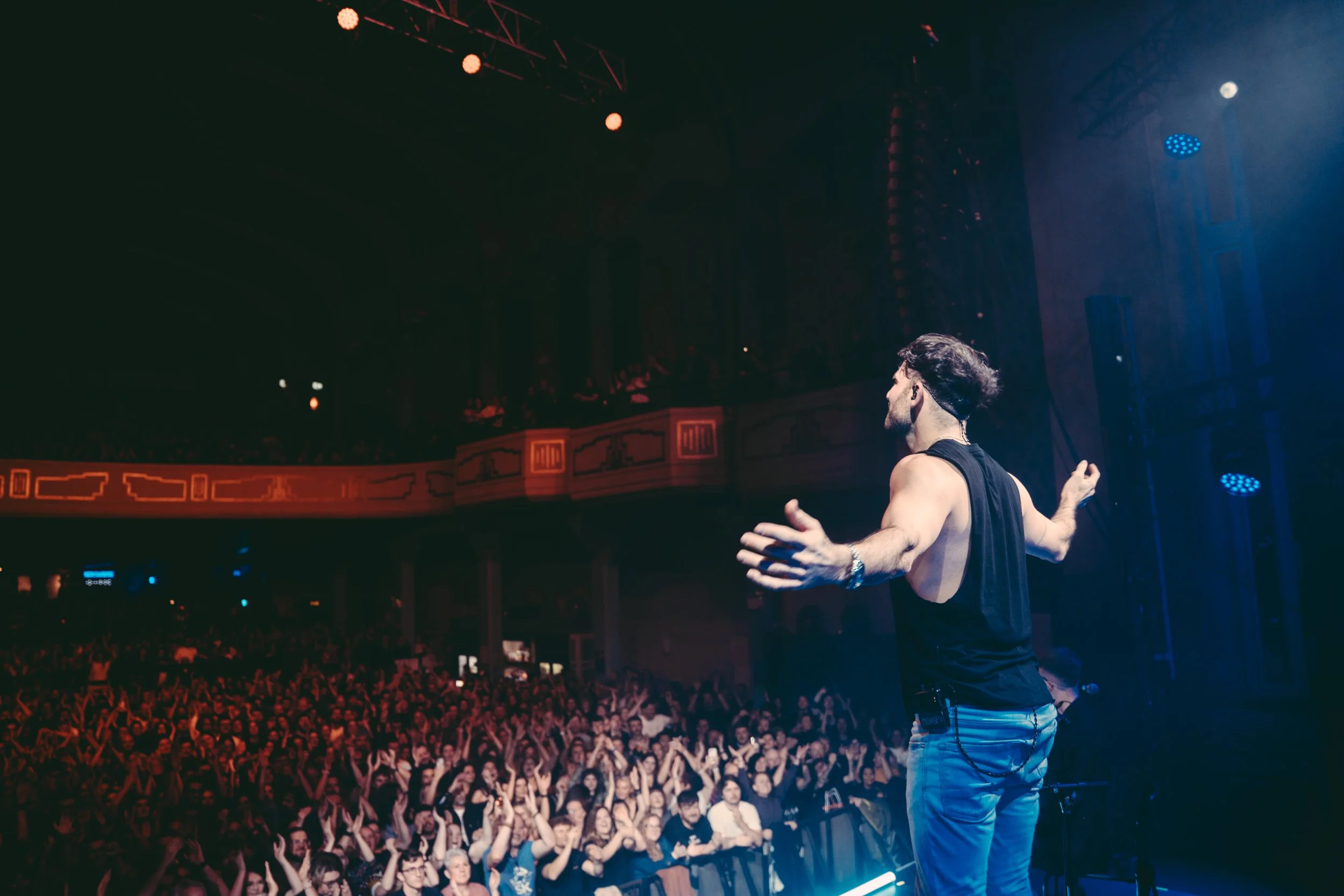 Performer on stage facing audience in a concert hall, with audience raising hands and cheering.