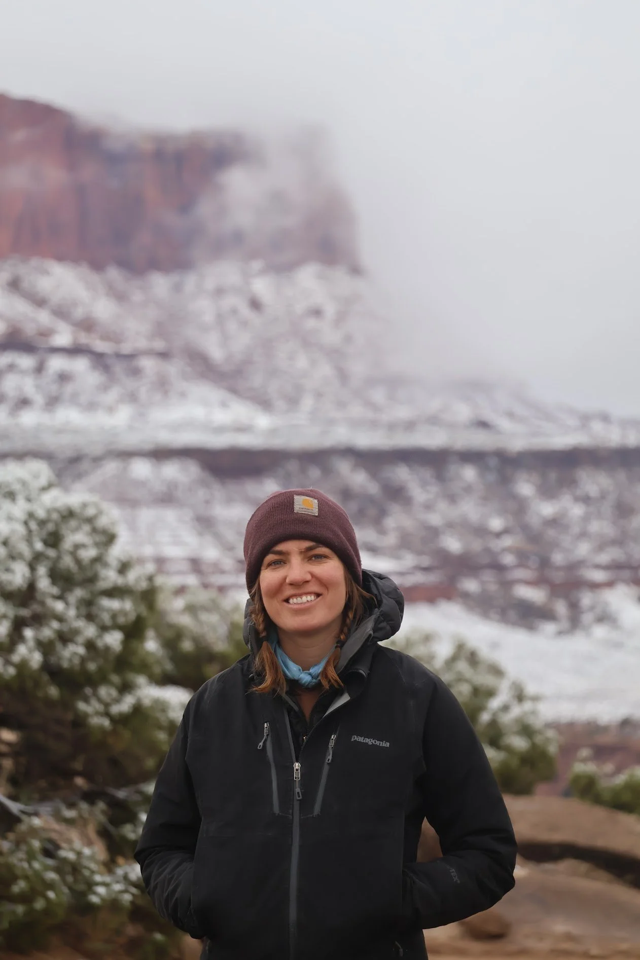 A woman smiling outdoors in a snowy, mountainous landscape, wearing a black Patagonia jacket and a brown Carhartt beanie.