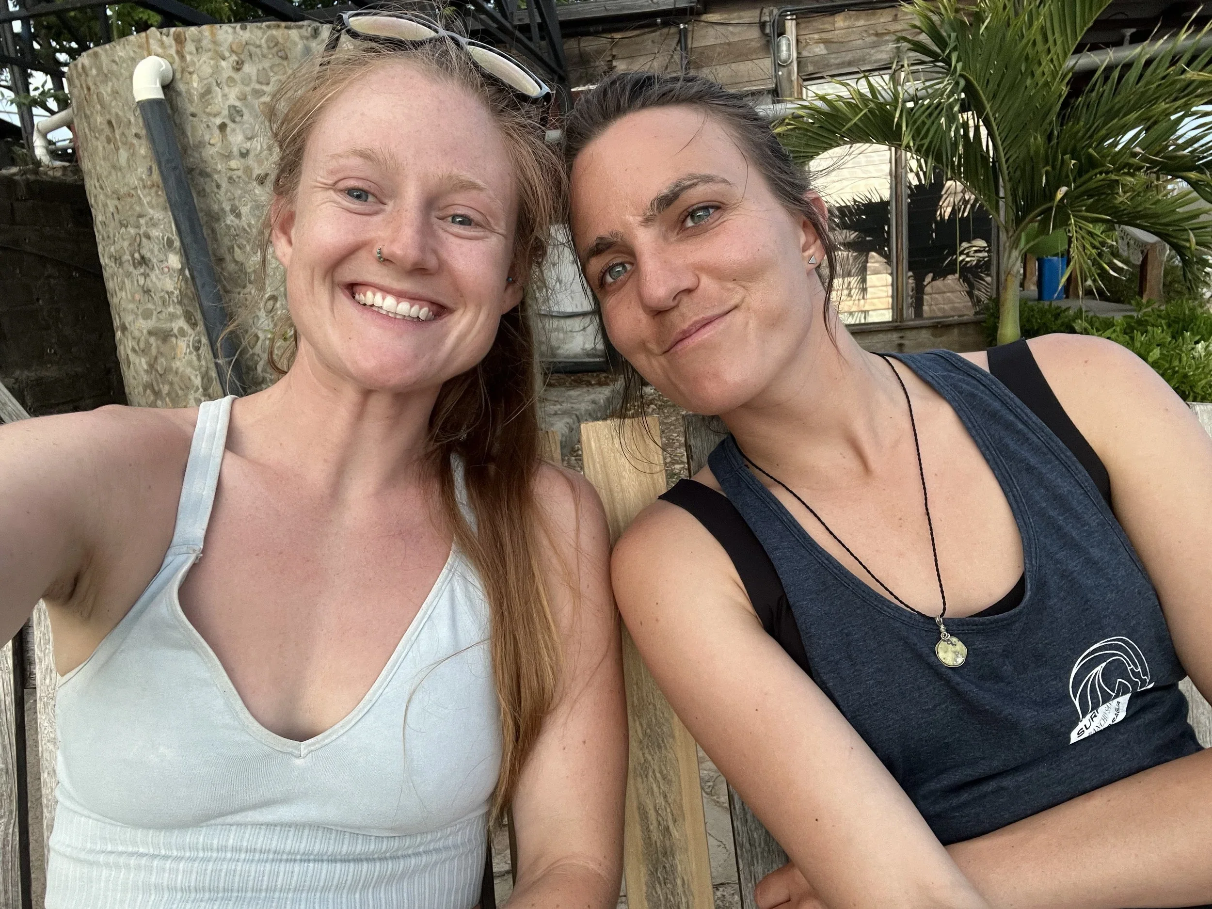 Two women smiling and taking a selfie outdoors, with plants and a wooden building in the background.