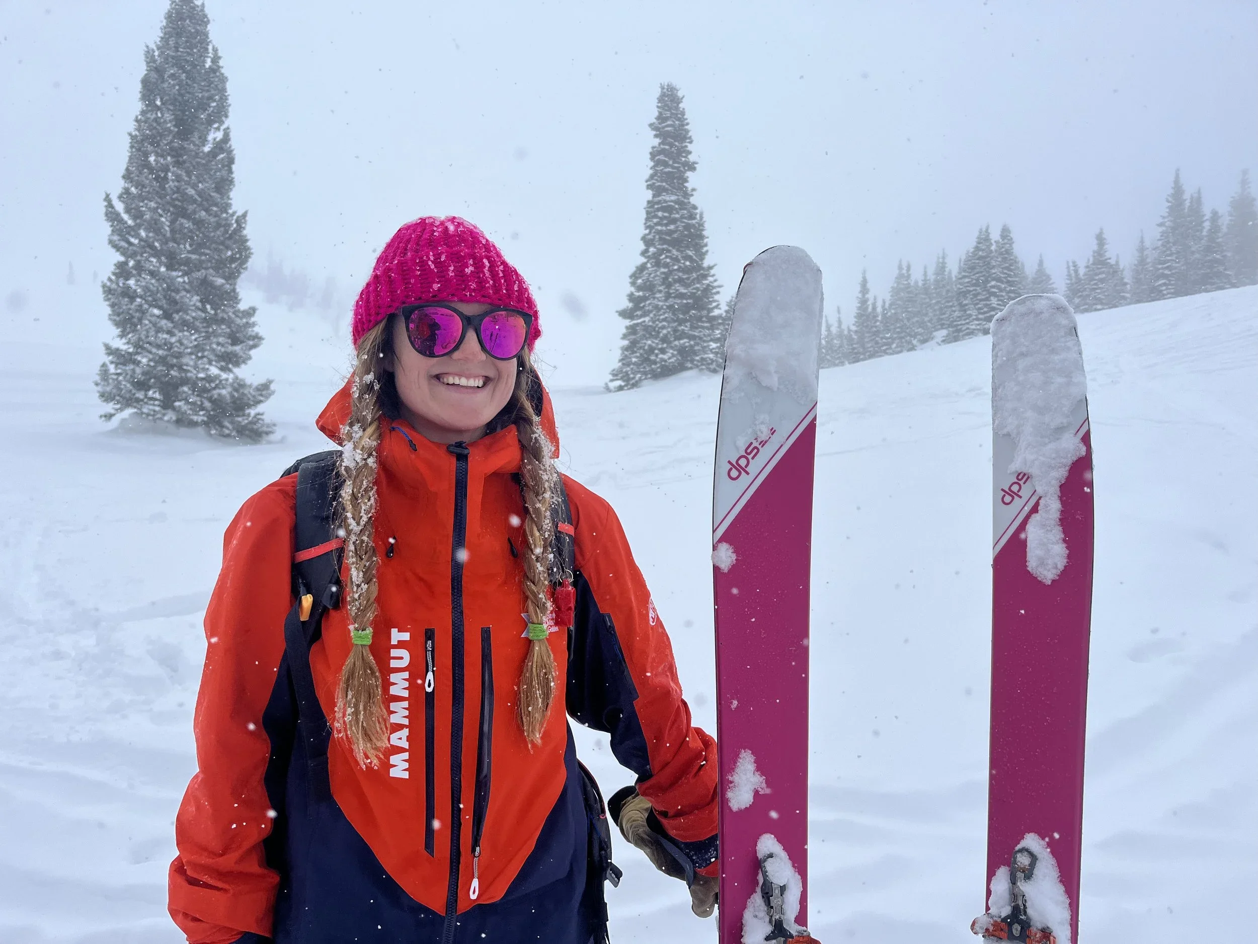 A woman in red ski gear, pink knit hat, and pink sunglasses standing in a snowy landscape with trees and holding pink skis.