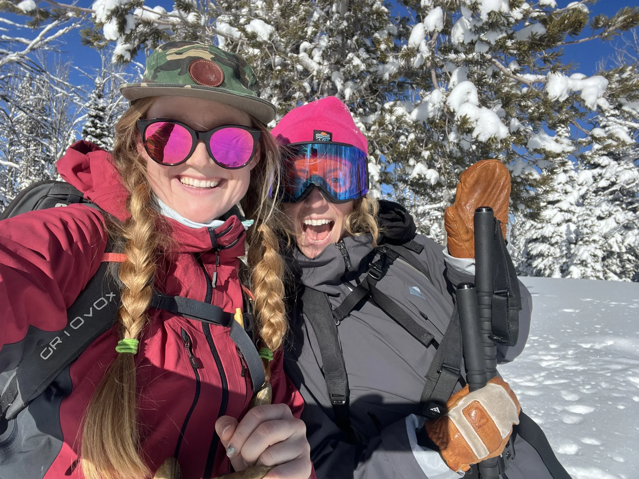 Two women in winter gear take a selfie in a snowy forest, smiling with snow-covered trees behind them.