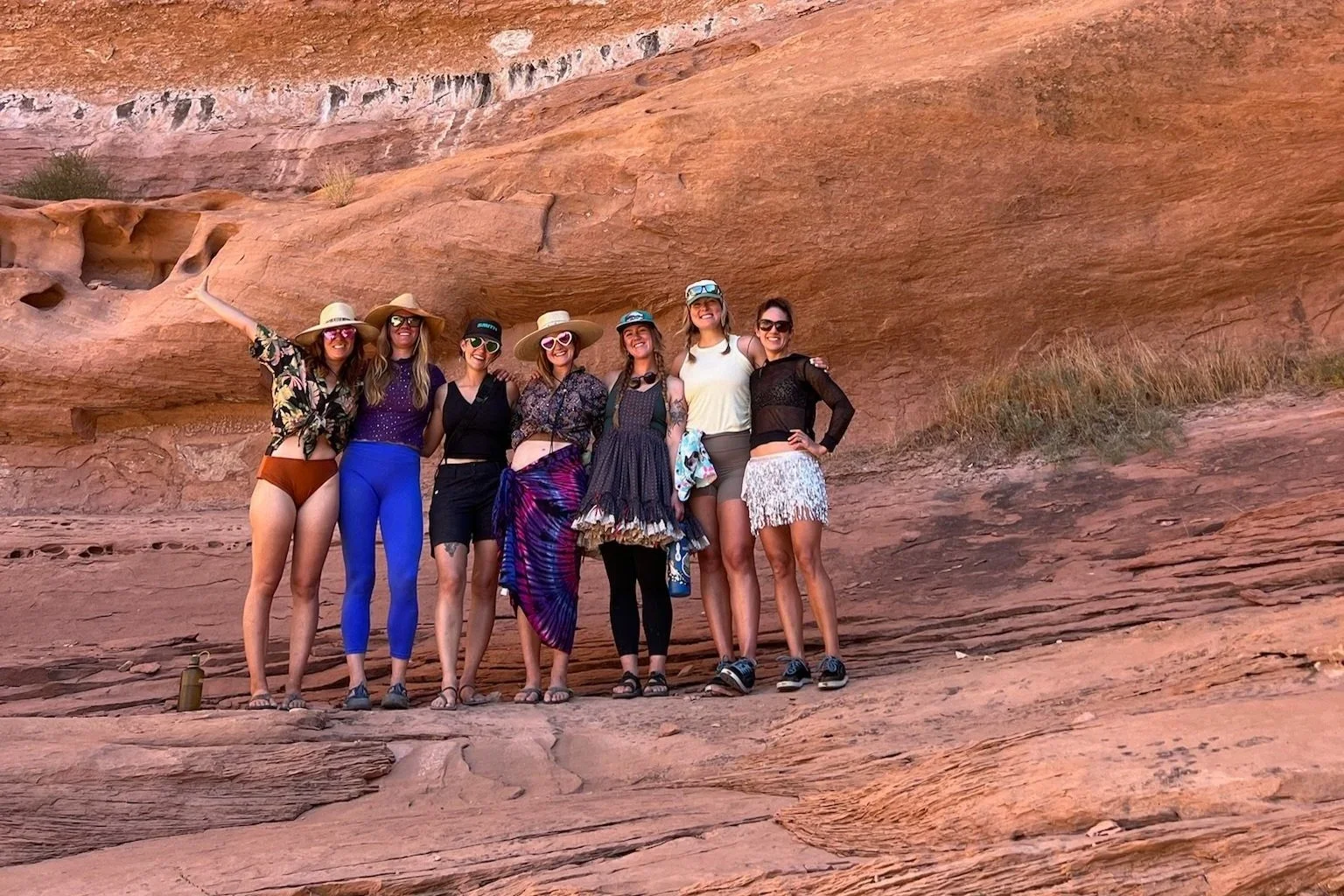 Group of seven women smiling and posing in front of red rock formation, wearing summer clothing, hats, and sunglasses.