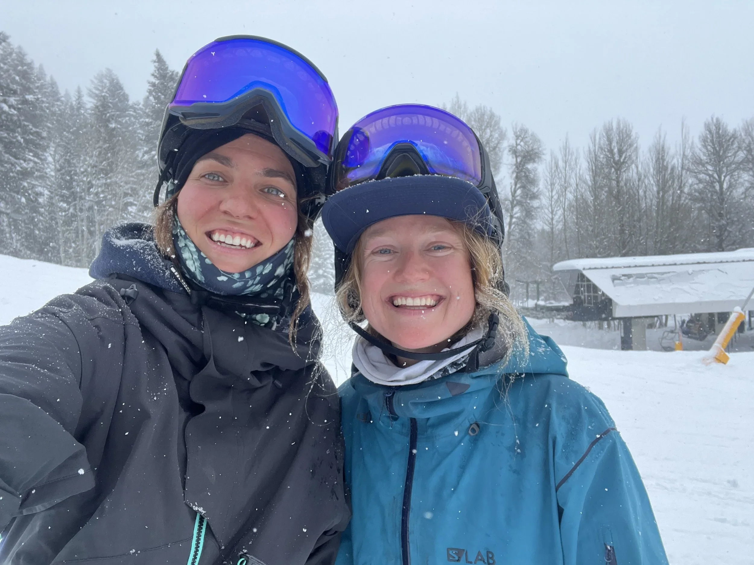 Two smiling women wearing ski helmets and goggles pose for a selfie in a snowy outdoor area with snow-covered trees and a small building in the background.
