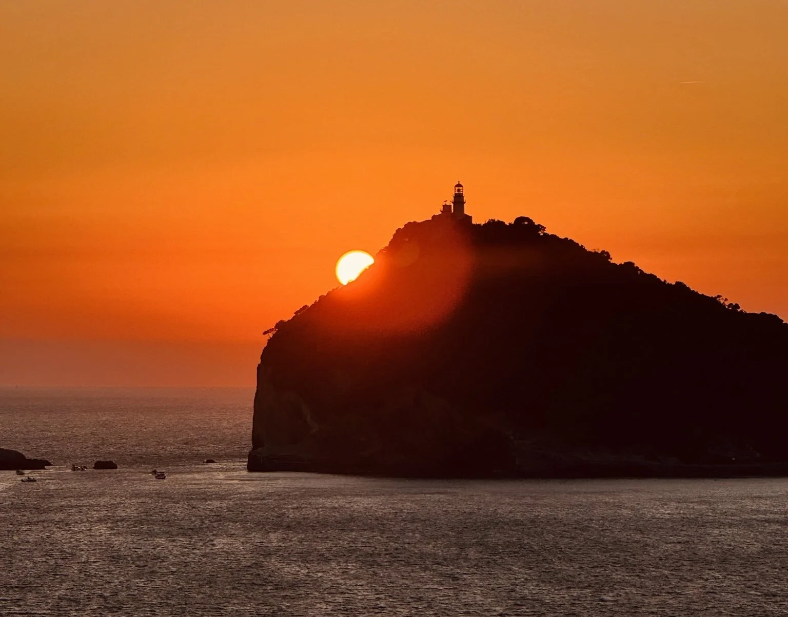 a sunset over the ocean peaking over an island with a lighthouse