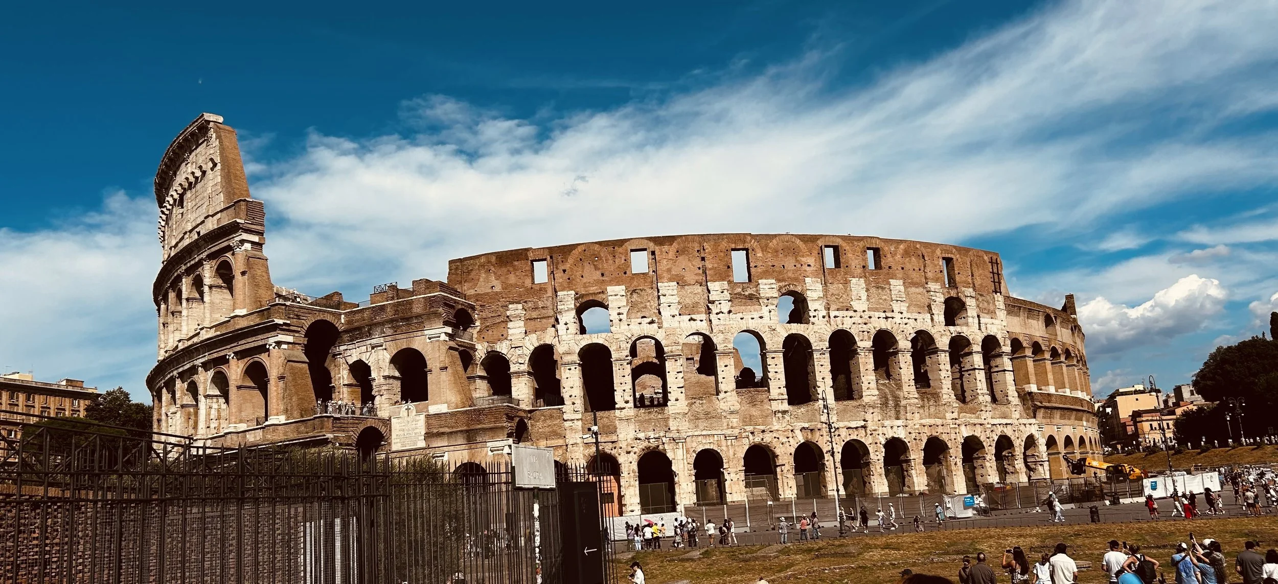 The Colosseum in Rome, Italy, seen under a blue sky with some clouds, with people walking around its perimeter.