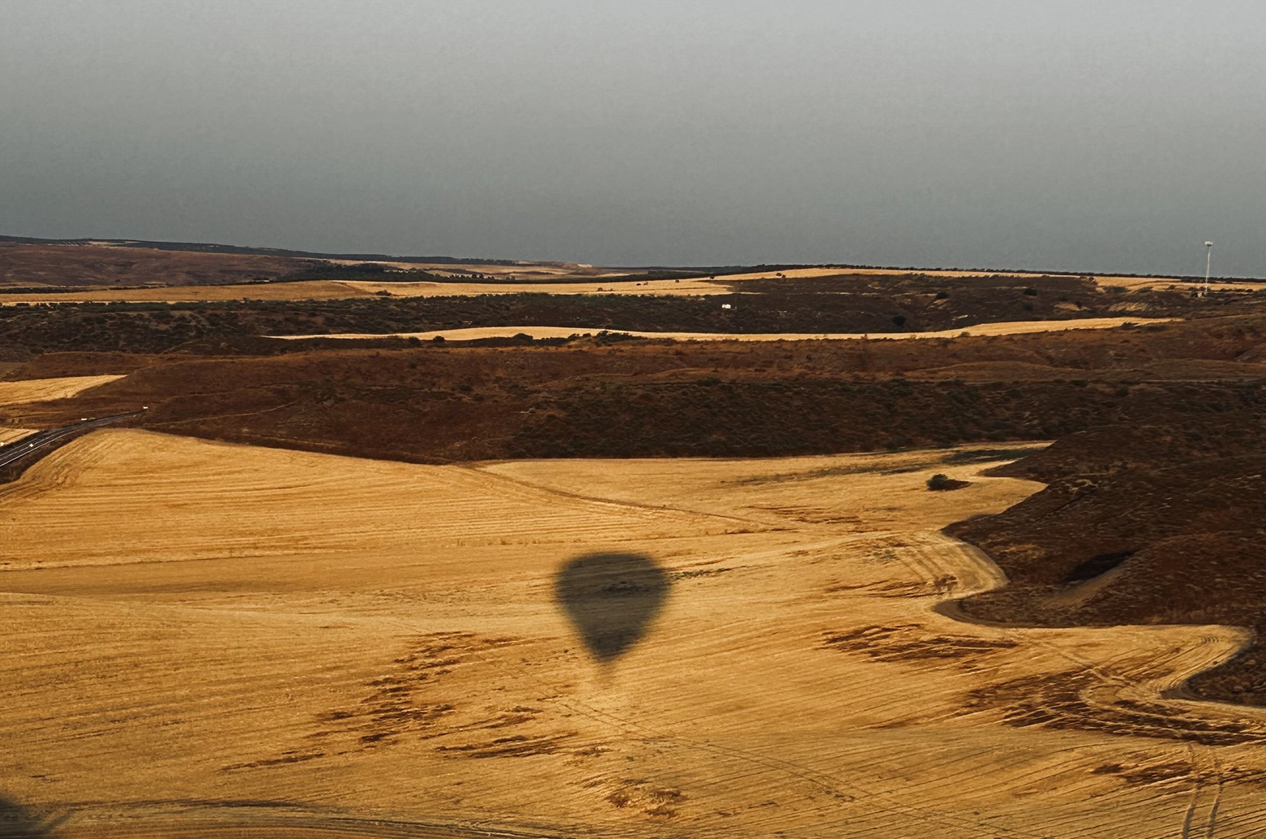 A landscape view of rolling hills with golden fields and patches of brown vegetation, and a large dark shadow of a hot air balloon cast on the ground, with a gray sky overhead.