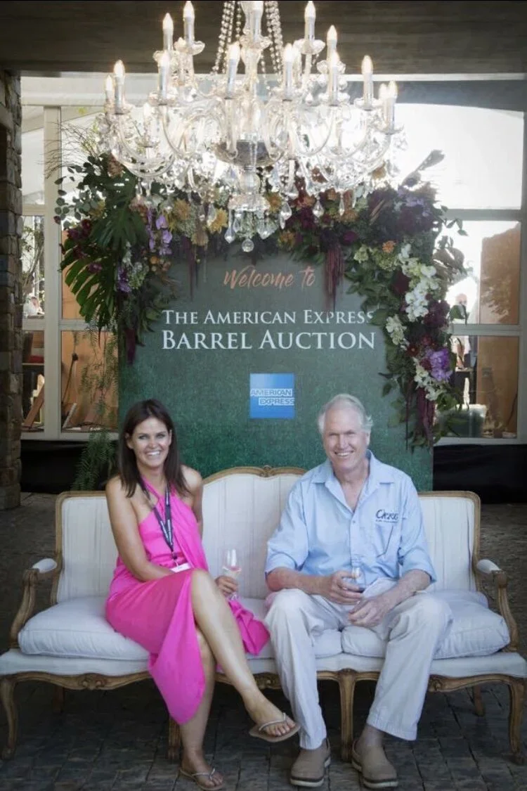 Shan and Matt Pascall sitting on a vintage white sofa holding glasses of Oneiric wine, at The American Express Barrel Auction.' 