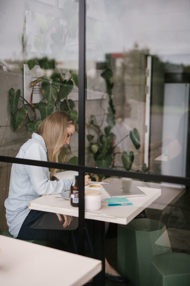 A Health and Wellness Copywriter sits by a window, working on a laptop with coffee, notebook, and lush plants nearby.