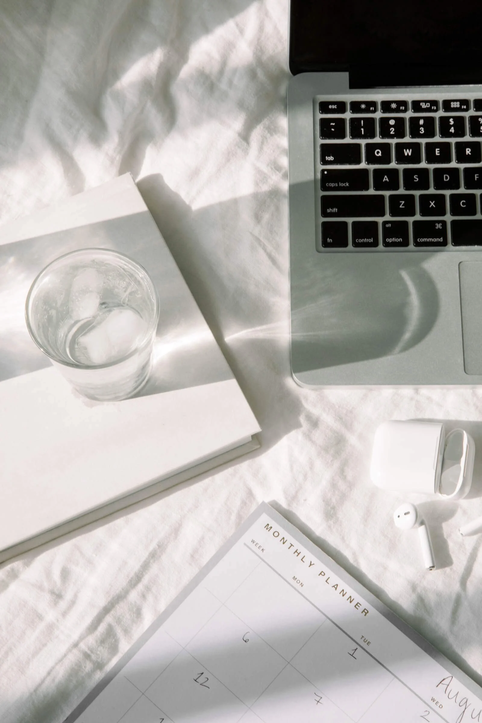 A glass of water, notebook, laptop, and planner on a bed in natural light.
