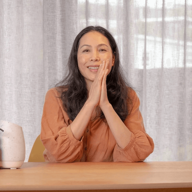 woman with long dark hair sitting at a table, smiling with clasped hands, in front of semi-transparent curtains