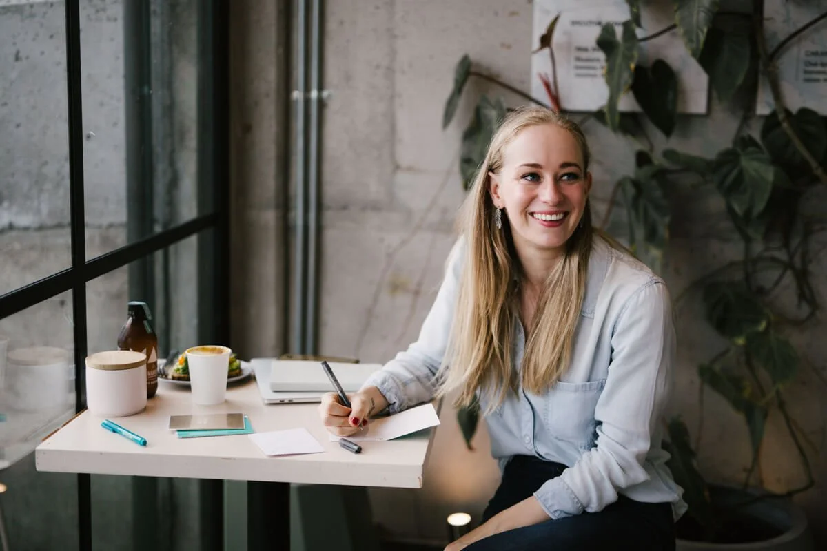 Smiling woman sits at a small table by a window, writing a card to a health and wellness client she created a website for, with a drink and plants nearby.