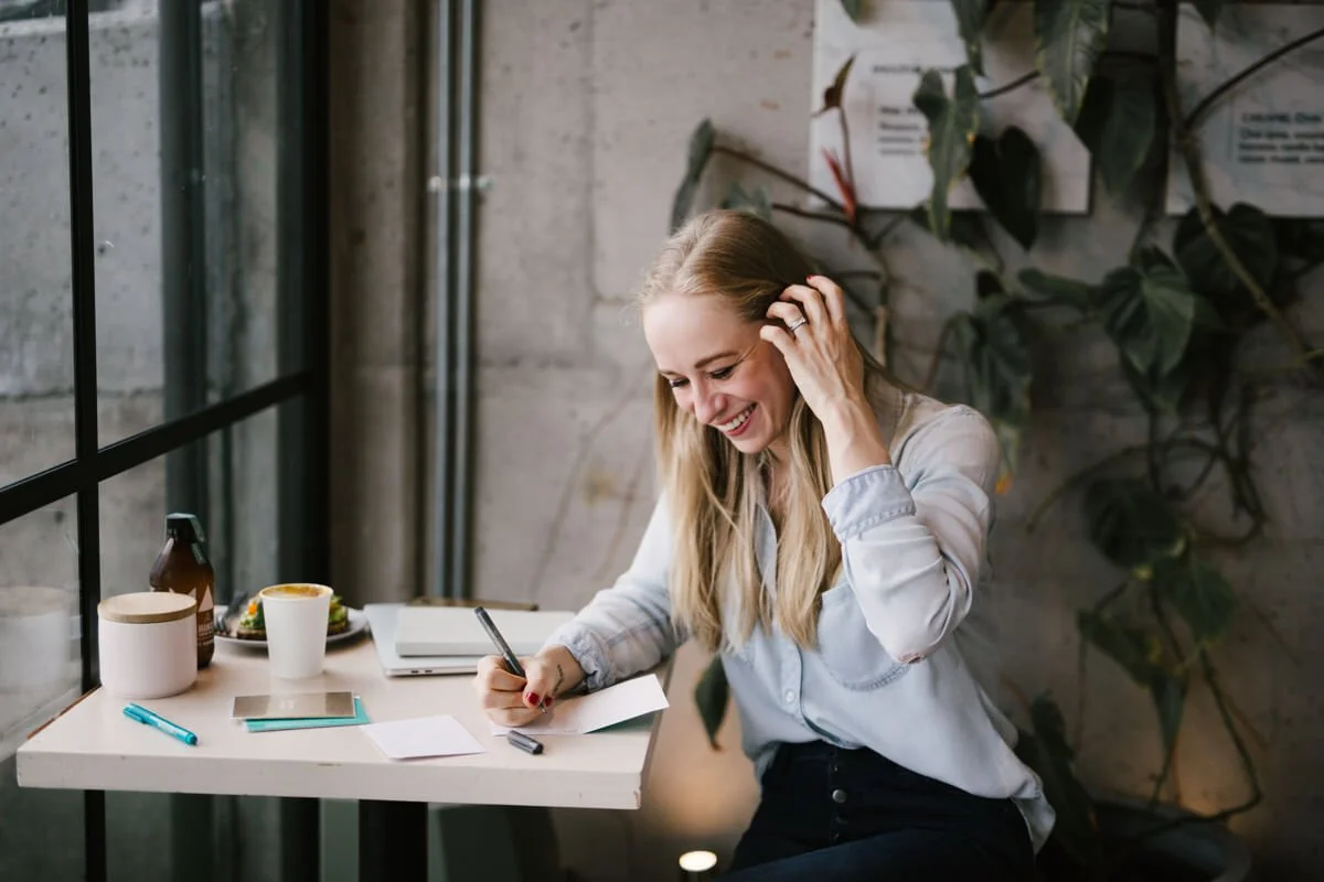A woman with long blonde hair, wearing a light-colored shirt, sitting at a table near a window, smiling while writing on a notepad with a pen. There are various items on the table, including a cup, a bottle, a plate of food, a smartphone, and some papers.