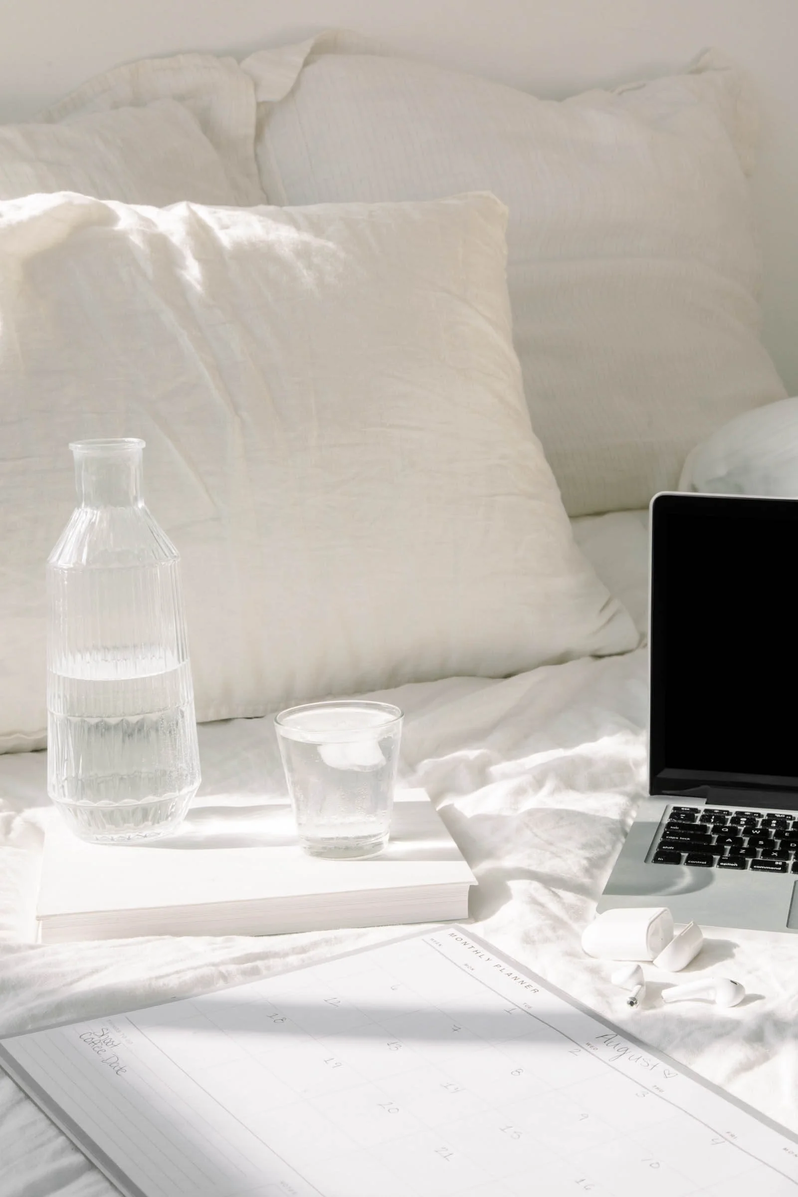 A glass carafe and cup of water, a laptop, and papers rest on a white bed with fluffy pillows in soft natural light.