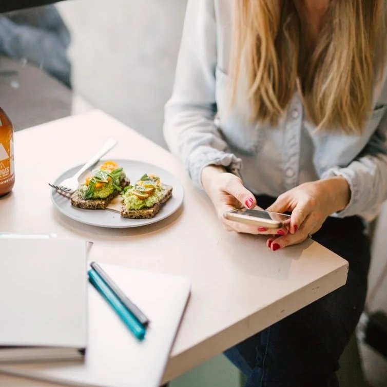 A woman with red nail polish using a smartphone at a table with avocado toast and a notebook with pens, emailing a wellness copywriting client from her phone