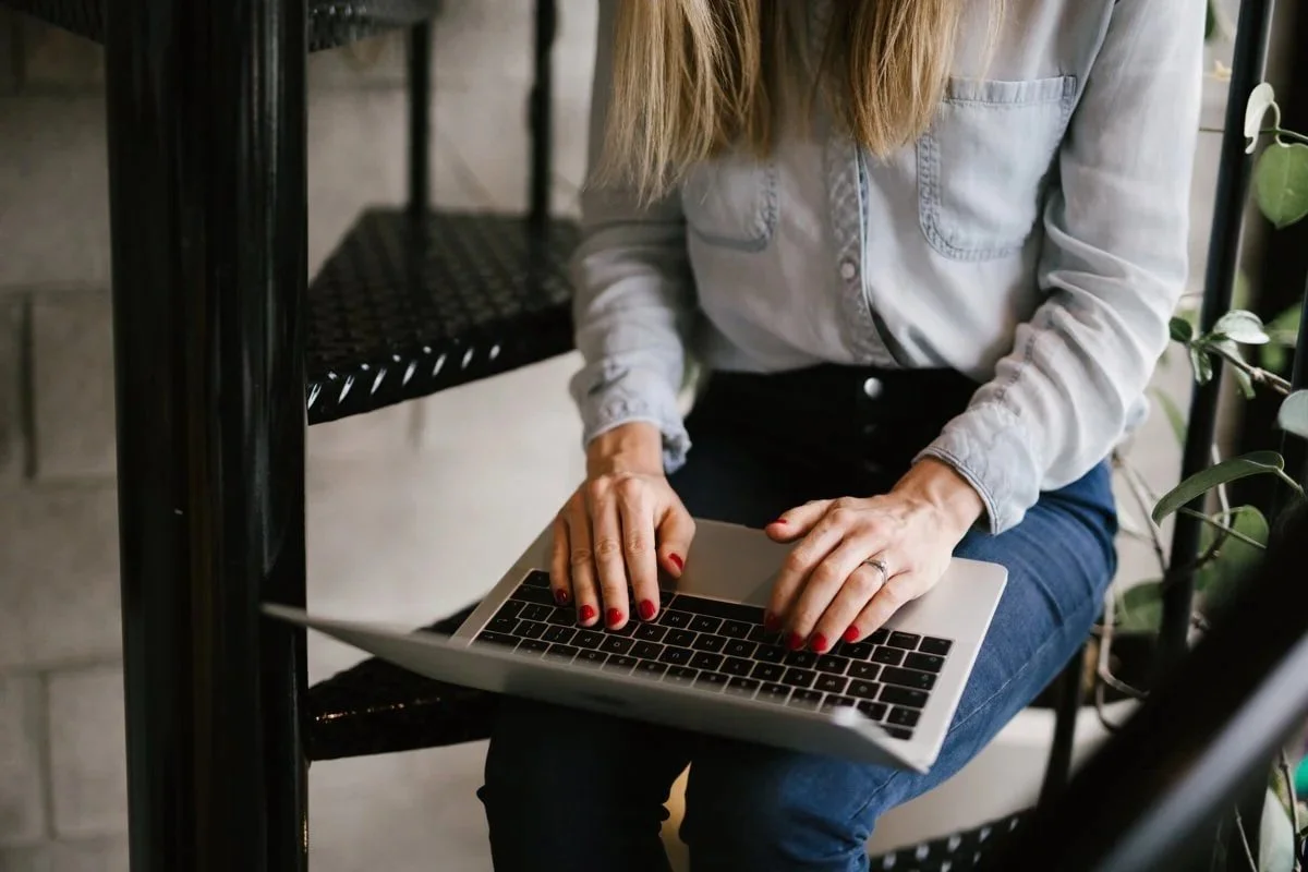 An Auckland NZ Squarespace website designer and copywriter with red nail polish types on a laptop while sitting on stairs next to a green plant.