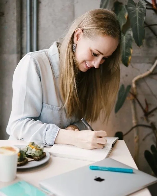 Female website copywriter with long blonde hair, wearing earrings and a light-colored shirt, is smiling while writing in a notebook at a table. There are some food items and a closed laptop on the table, with a plant and a concrete wall.