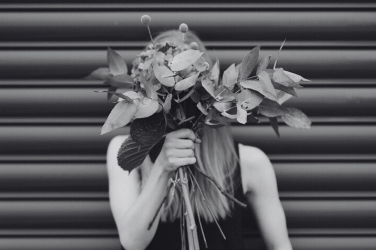 A wellness copywriter holding a bouquet of leaves and flowers covers their face before a striped metal background.