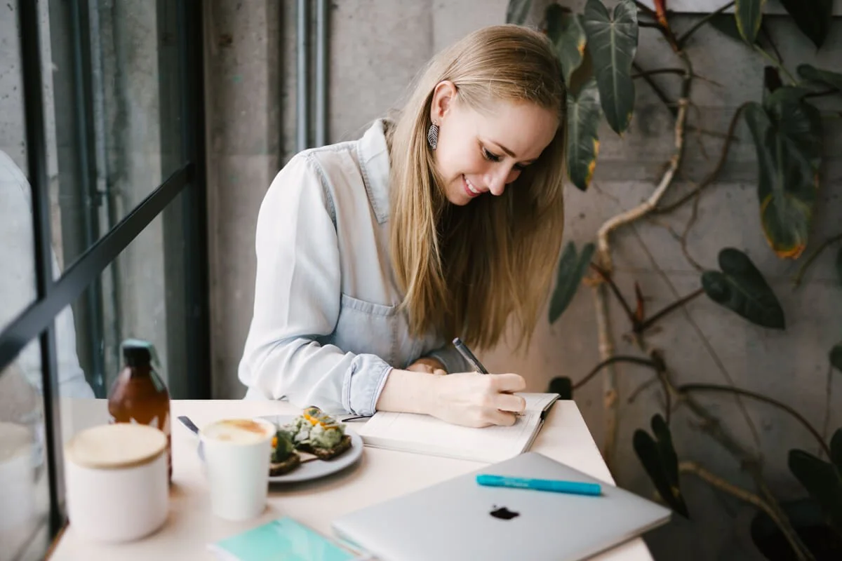 Auckland Copywriter and Website Designer sits working at desk in cafe with laptop, avocado toast and tumeric latte nearby