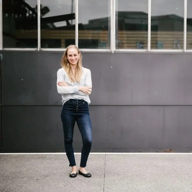 A young woman with long blonde hair, wearing a light gray shirt and dark jeans, smiling with her arms crossed, standing outdoors in front of a dark gray wall with large windows.
