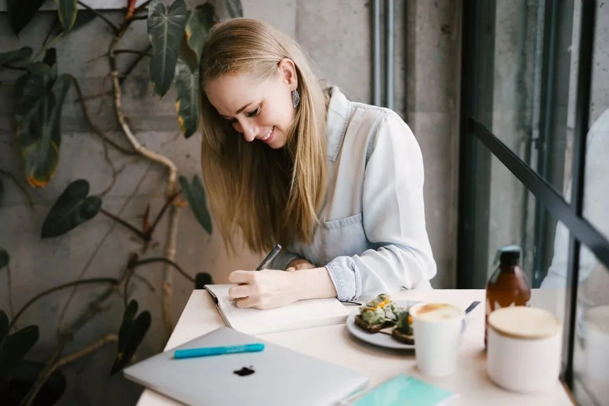 A woman with long blonde hair sitting at a table, smiling, writing in a notebook, with a laptop, a cup of coffee, a plate of food, and a small bottle nearby, in front of large windows and a green leafy plant.