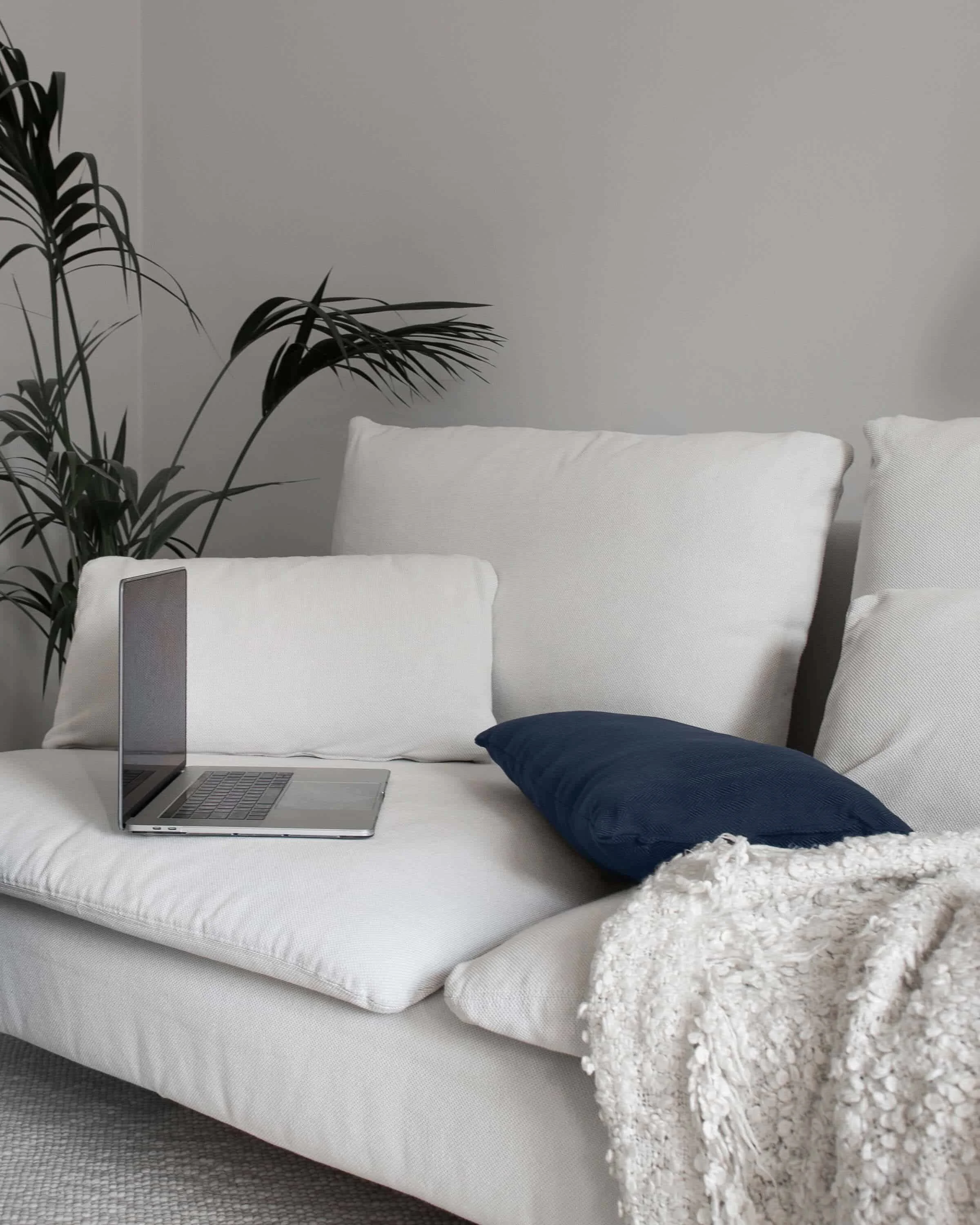 A silver laptop and navy pillow rest on a white sofa with a textured blanket, creating a perfect wellness copywriting nook.